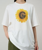 Model wearing a Gramicci white G-Flower Tee with sunflower graphic against a light tone background.
