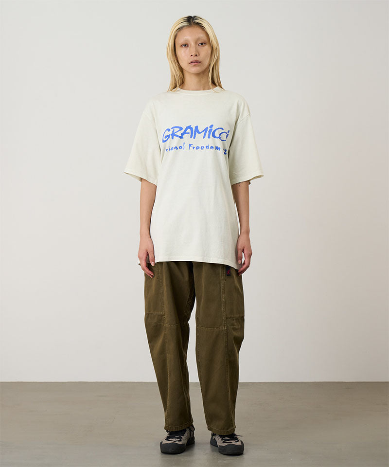 Model wearing a natural pigment Gramicci Freedom Tee Pigment Dyed with a blue graphic and brown pants on a light tone background.