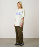 Model wearing a natural pigment Gramicci Freedom Tee Pigment Dyed with a blue graphic and brown pants on a light tone background.