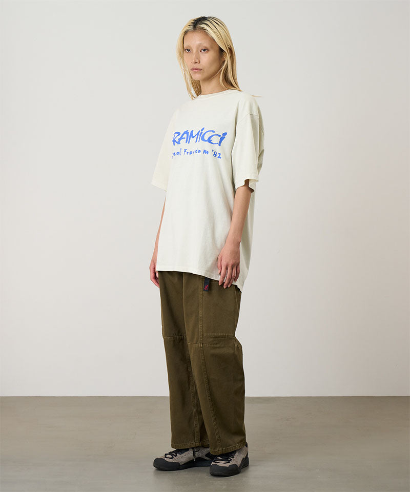 Model wearing a natural pigment Gramicci Freedom Tee Pigment Dyed with a blue graphic and brown pants on a light tone background.