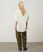 Back view of a model wearing a natural pigment Gramicci Freedom Tee Pigment Dyed and brown pants on a light tone background.