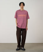 Model wearing a red pigment Gramicci Freedom Tee Pigment Dyed with a mango colored graphic and brown pants on a light tone background.