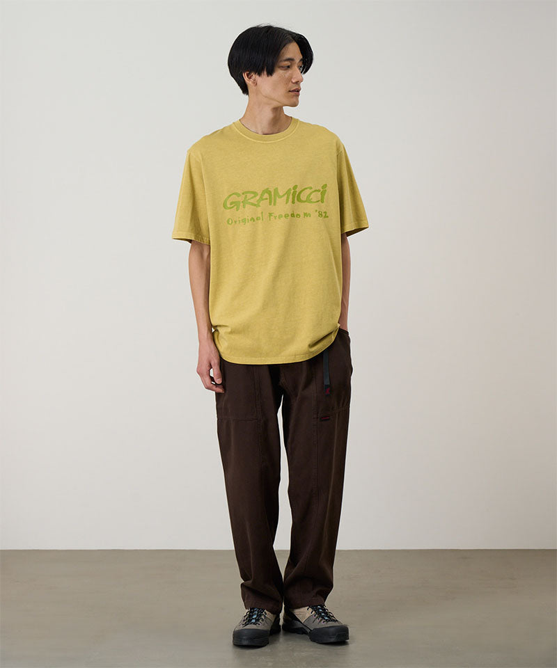 Model wearing a yellow sulpher pigment Gramicci Freedom Tee Pigment Dyed with a lime green graphic and brown pants on a light tone background.