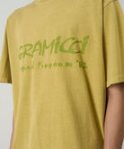 Model wearing a Gramicci yellow sulpher pigment Freedom Tee Pigment Dyed with lime green graphic against a light tone background.