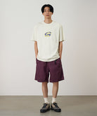 Model wearing a natural colored Gramicci G-Scribble Tee Pigment Dyed shirt with a purple graphic and purple shorts on a light tone background.
