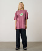 Model wearing a red Gramicci G-Scribble Tee Pigment Dyed shirt with a green graphic and green plaid shorts on a light tone background.
