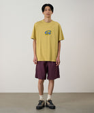 Model wearing a sulpher Gramicci G-Scribble Tee Pigment Dyed shirt with a blue graphic and purple shorts on a light tone background.
