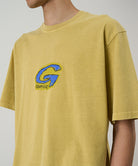 Model wearing a Gramicci sulpher G-Scribble Tee Pigment Dyed shirt with blue graphic against a light tone background.
