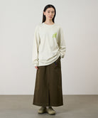 Model wearing a natural pigment Gramicci Running Man L/S Tee Pigment Dyed with a lime green graphic and brown skirt on a light-tone background.