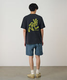 Person wearing a black t-shirt with green graphics and plaid shorts on a plain background