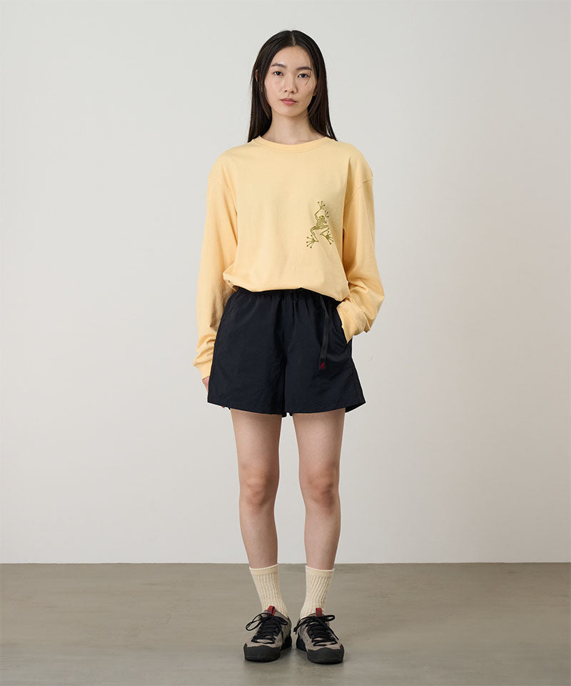 Model wearing a mango colored Gramicci Sticky Hands L/S Tee with a green frog graphic and black shorts on a light tone background.