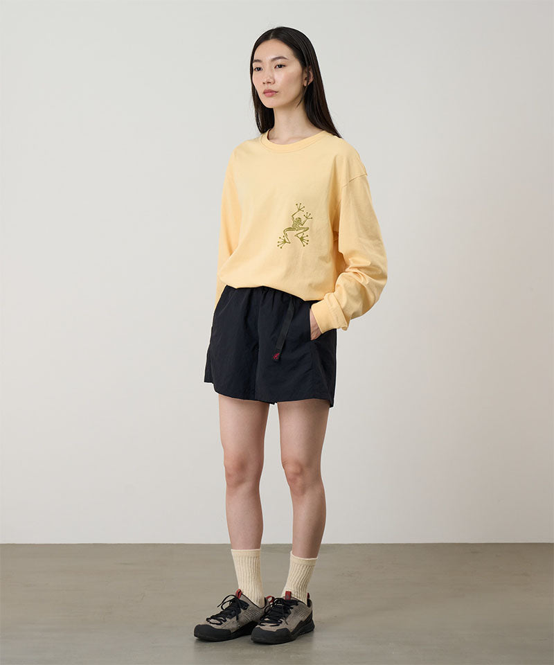 Model wearing a mango colored Gramicci Sticky Hands L/S Tee with a green frog graphic and black shorts on a light tone background.