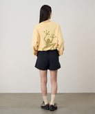 Rear view of a model wearing a mango colored Gramicci Sticky Hands L/S Tee with a green frog graphic and black shorts on a light tone background.
