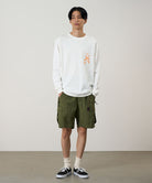 Model wearing a white Gramicci Sticky Hands L/S Tee with a orange frog graphic and olive shorts on a light tone background.
