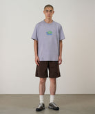 Model wearing a lavender Gramicci Turtle Tee with a a turtle graphic and brown shorts on a light tone background.
