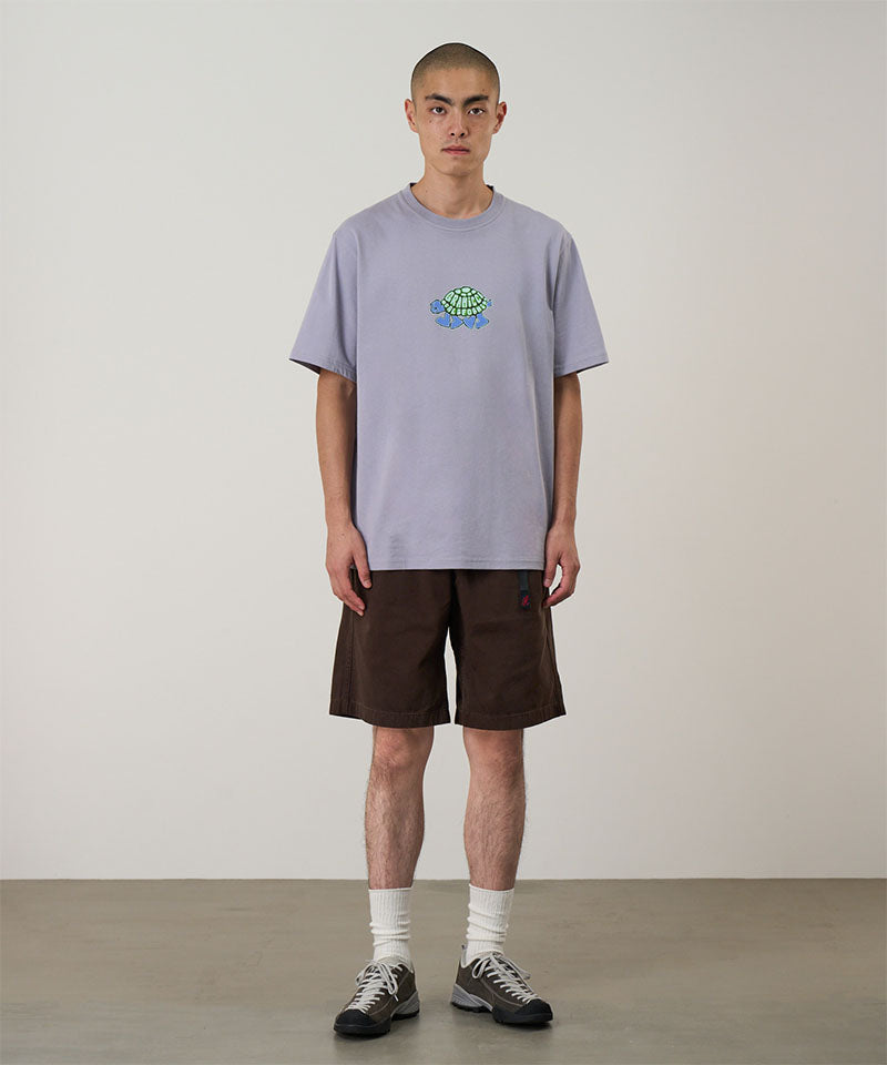 Model wearing a lavender Gramicci Turtle Tee with a a turtle graphic and brown shorts on a light tone background.
