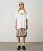 Model wearing a white Gramicci Turtle Tee with a a turtle graphic and beige skirt on a light tone background.
