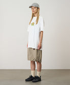 Model wearing a white Gramicci Turtle Tee with a a turtle graphic and beige skirt on a light tone background.
