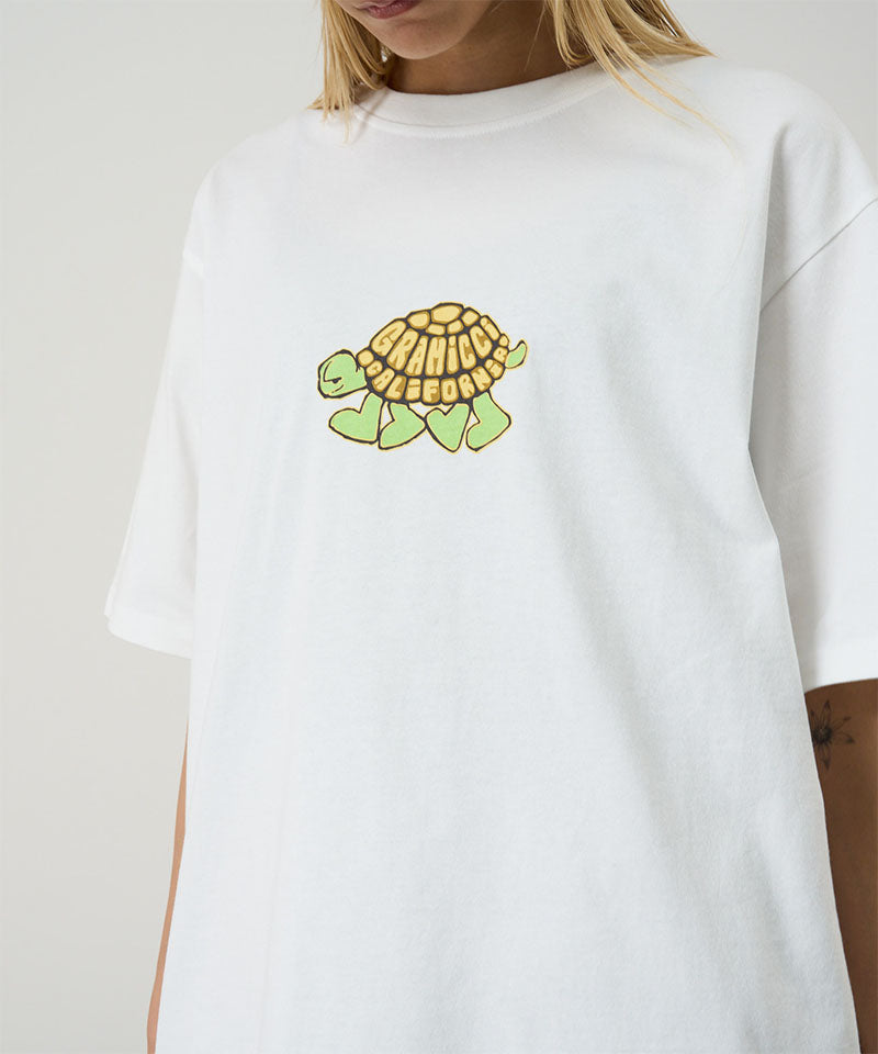 Model wearing a Gramicci white Turtle Tee with a turtle graphic against a light tone background.
