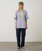 Model wearing a lavender Gramicci Quencher Tee with a hydration drink graphic and blue pants on a light tone background.