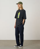 Model wearing a vintage black Gramicci Quencher Tee with a hydration drink graphic and blue pants on a light tone background.