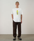 Model wearing a white Gramicci Quencher Tee with a hydration drink graphic and brown pants on a light tone background.