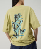 Back view of a model wearing a Gramicci lime colored Ice Sculpture Tee with a running man graphic against a light tone background.