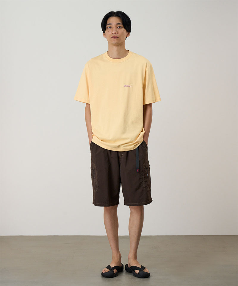 Model wearing a mango colored Gramicci Ice Sculpture Tee with a a running man graphic and brown shorts on a light tone background.