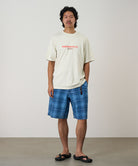 Model wearing a natural pigment Gramicci Footprints Tee Pigment Dyed with a red preserve it graphic and blue plaid shorts on a light tone background.