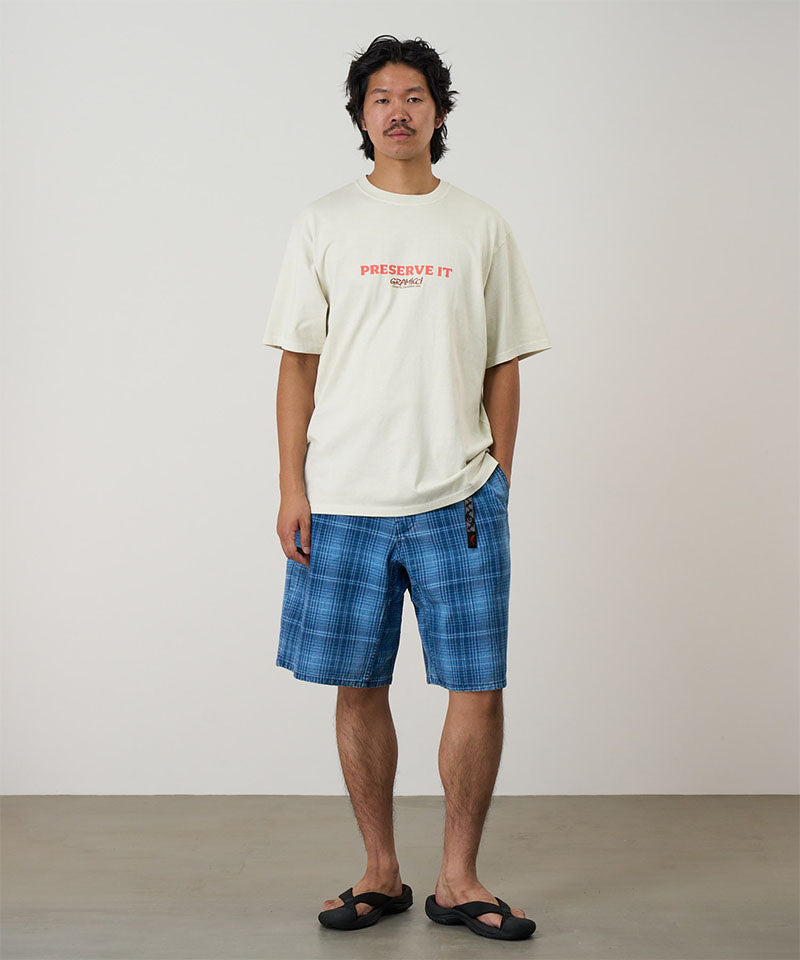 Model wearing a natural pigment Gramicci Footprints Tee Pigment Dyed with a red preserve it graphic and blue plaid shorts on a light tone background.