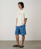 Model wearing a natural pigment Gramicci Footprints Tee Pigment Dyed with a red preserve it graphic and blue plaid shorts on a light tone background.