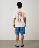 Rear view of a model wearing a natural pigment Gramicci Footprints Tee Pigment Dyed with a red graphic and blue plaid shorts on a light tone background.