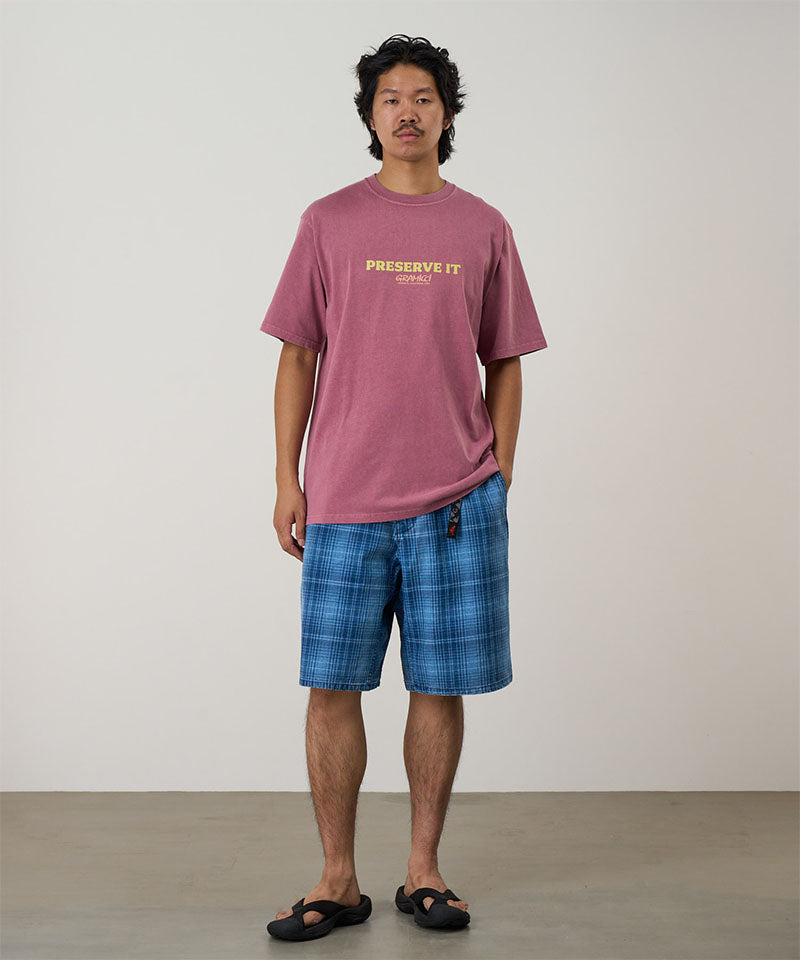 Model wearing a red pigment Gramicci Footprints Tee Pigment Dyed with a yellow graphic and blue plaid shorts on a light tone background.