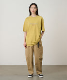 Model wearing a sulphur pigment Gramicci Footprints Tee Pigment Dyed with a grey graphic and beige pants on a light tone background.