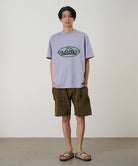 Model wearing a lavender Gramicci Half Dome Tee with a grey and green graphic and green shorts on a light tone background.
