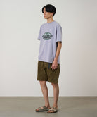 Model wearing a lavender Gramicci Half Dome Tee with a grey and green graphic and green shorts on a light tone background.
