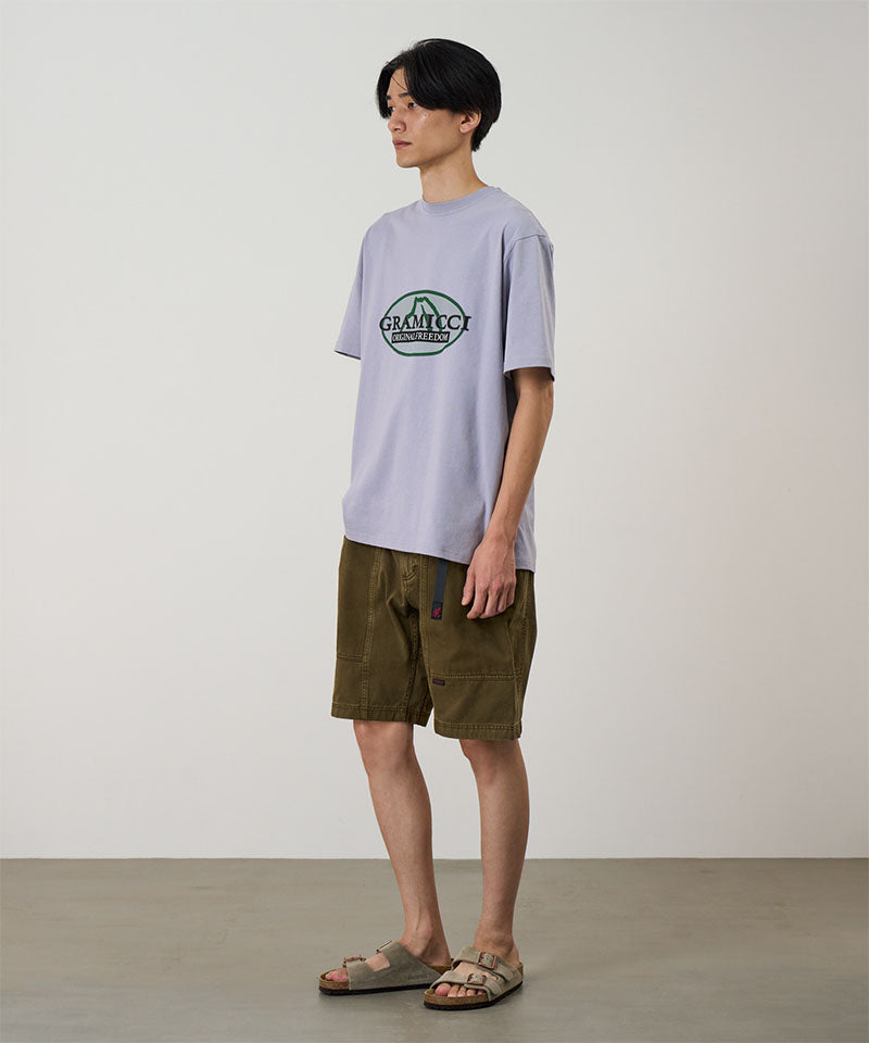 Model wearing a lavender Gramicci Half Dome Tee with a grey and green graphic and green shorts on a light tone background.
