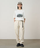 Model wearing a white Gramicci Half Dome Tee with a grey and green graphic and light beige pants on a light tone background.
