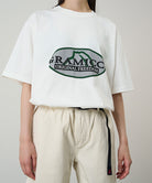 Model wearing a white Gramicci Half Dome Tee with a grey and green graphic and light beige pants on a light tone background.
