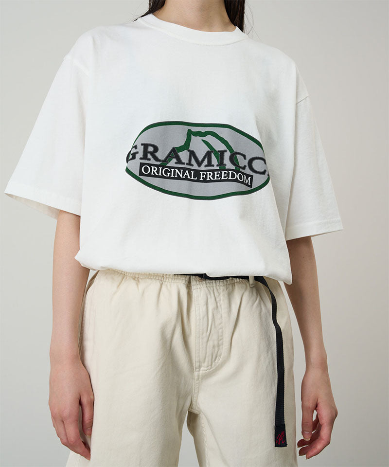 Model wearing a white Gramicci Half Dome Tee with a grey and green graphic and light beige pants on a light tone background.
