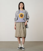 Person wearing a plaid shirt over a graphic t-shirt with a sunflower design, beige skirt, and black sneakers on a plain background.