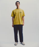 Model wearing a yellow Gramicci G-Sail Tee with black graphic and blue pants on a light-tone background.