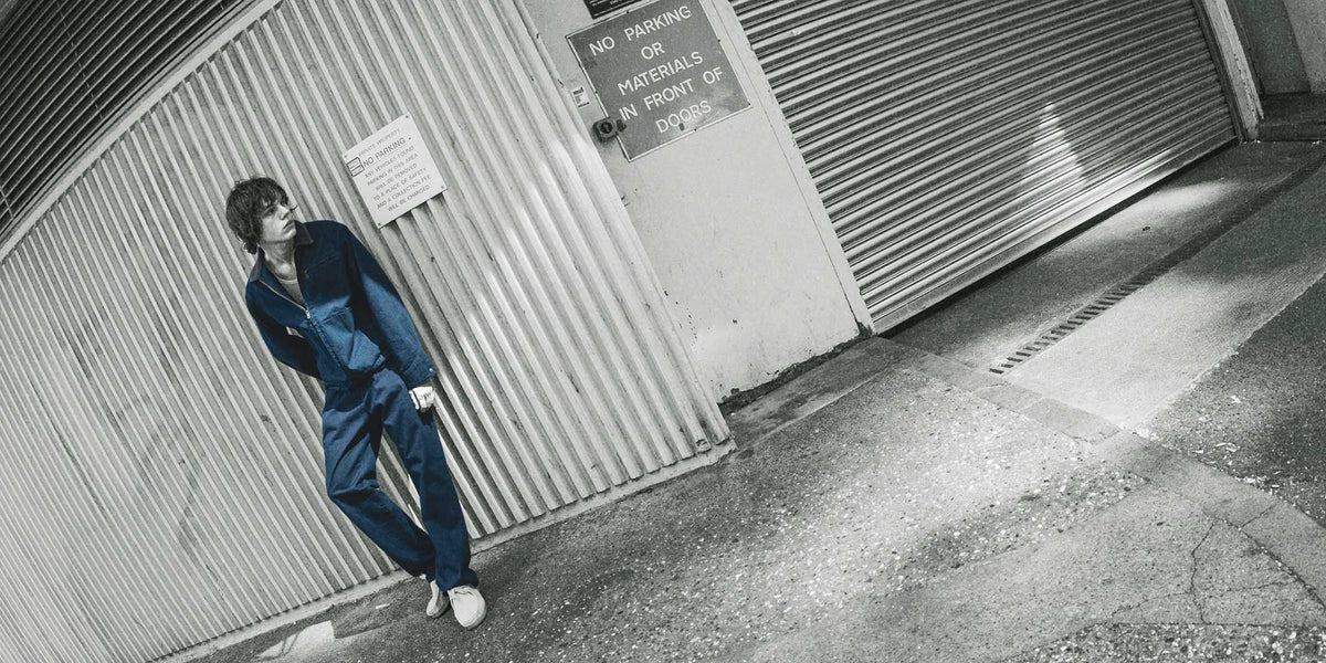 Gramicci relaxed-fit blue denim jacket and matching pants worn by a person leaning against a corrugated metal wall in an industrial alleyway.