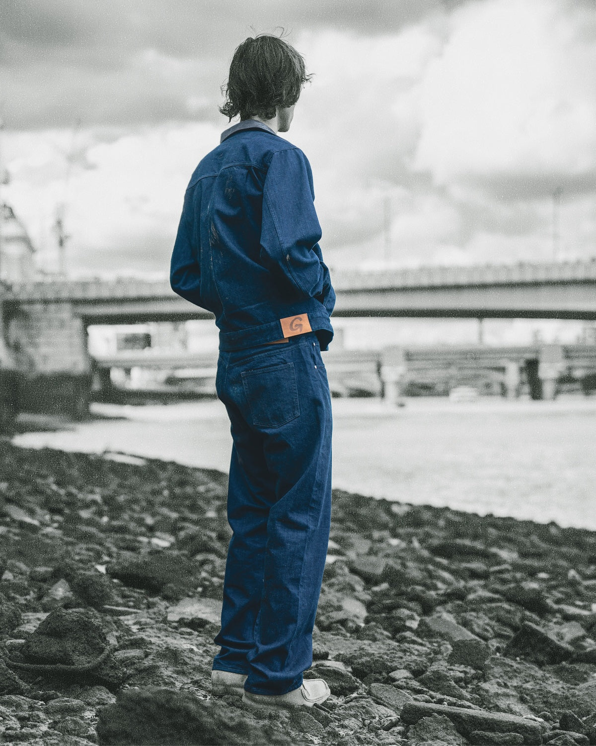 Gramicci blue denim jacket and matching relaxed pants worn by a person standing on a rocky shore overlooking the water and a bridge.   