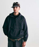 Model wearing a Gramicci black hooded half-zip anorak layered over a plaid shirt, dark corduroy pants, and a fleece brimmed cap with sunglasses.   