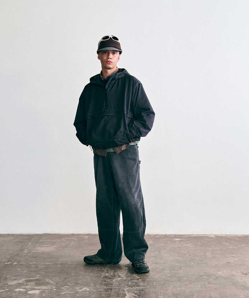 Model wearing a Gramicci black hooded half-zip anorak, dark corduroy pants, dark trail shoes, and a fleece brimmed cap with sunglasses.   