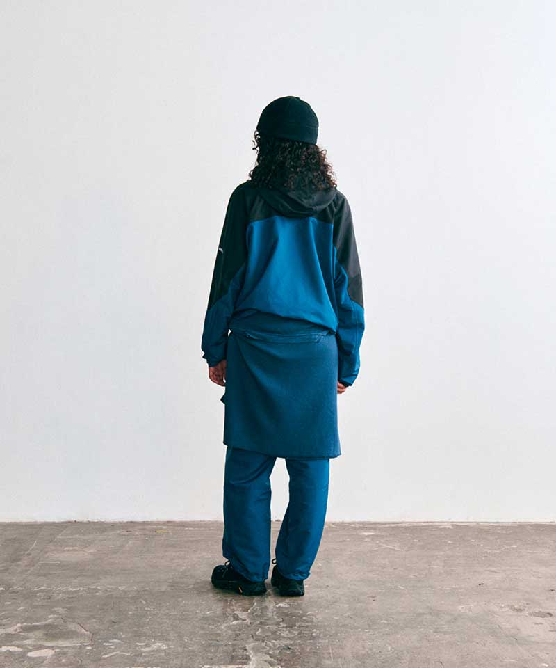 Model wearing a Gramicci teal and black colorblock hooded jacket, matching teal pants with a sweatshirt tied around the waist, a black beanie, and black trail shoes, seen from behind.   
