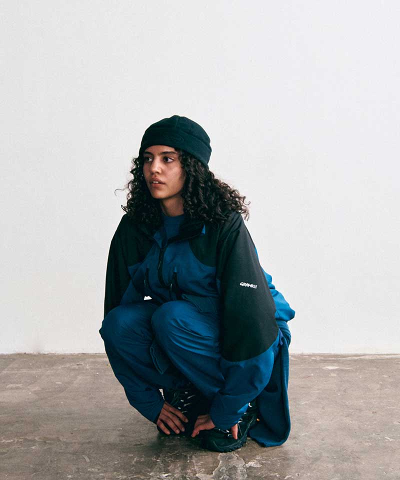 Model wearing a Gramicci teal and black colorblock zip-up jacket, matching teal pants, a black beanie, and black trail shoes.   