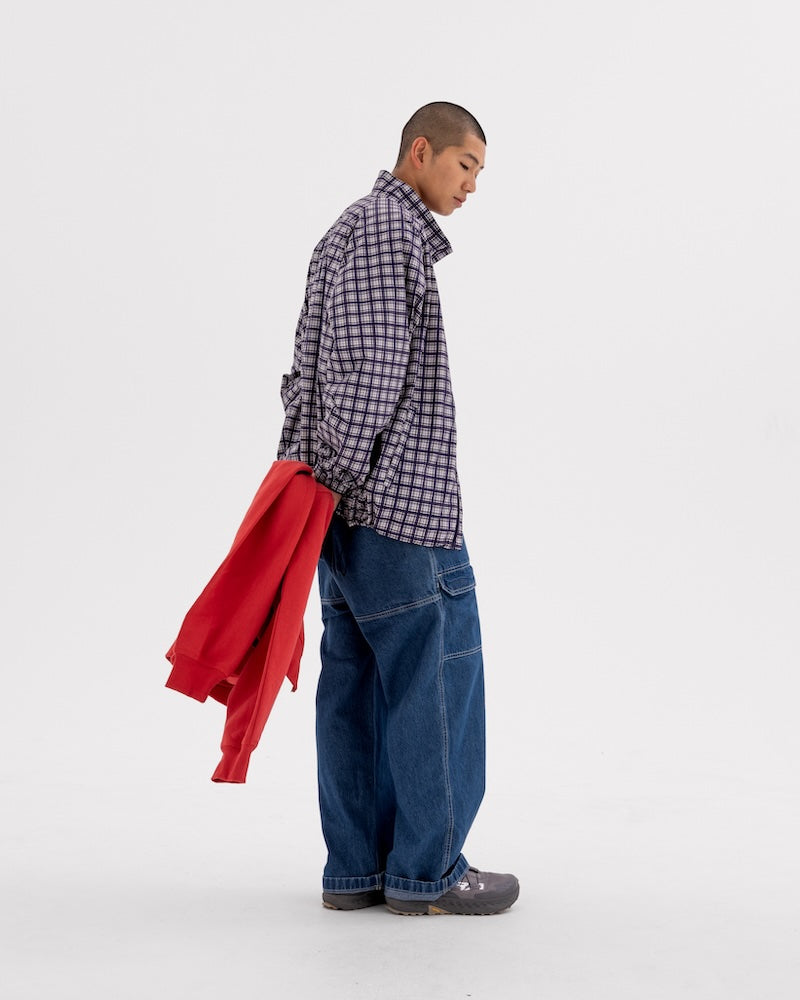 Model holding a red jacket, wearing a checkered button down Gramicci shirt and jeans, on a light-tone background.