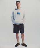 Model wearing the Gramicci Blueberry Hooded Sweatshirt with blueberry graphic in heather ash and navy colored shorts on a light tone background.
